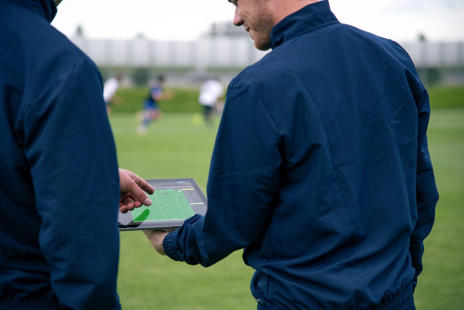 Coaching staff using KINEXON sports analytics software on a tablet beside a football pitch